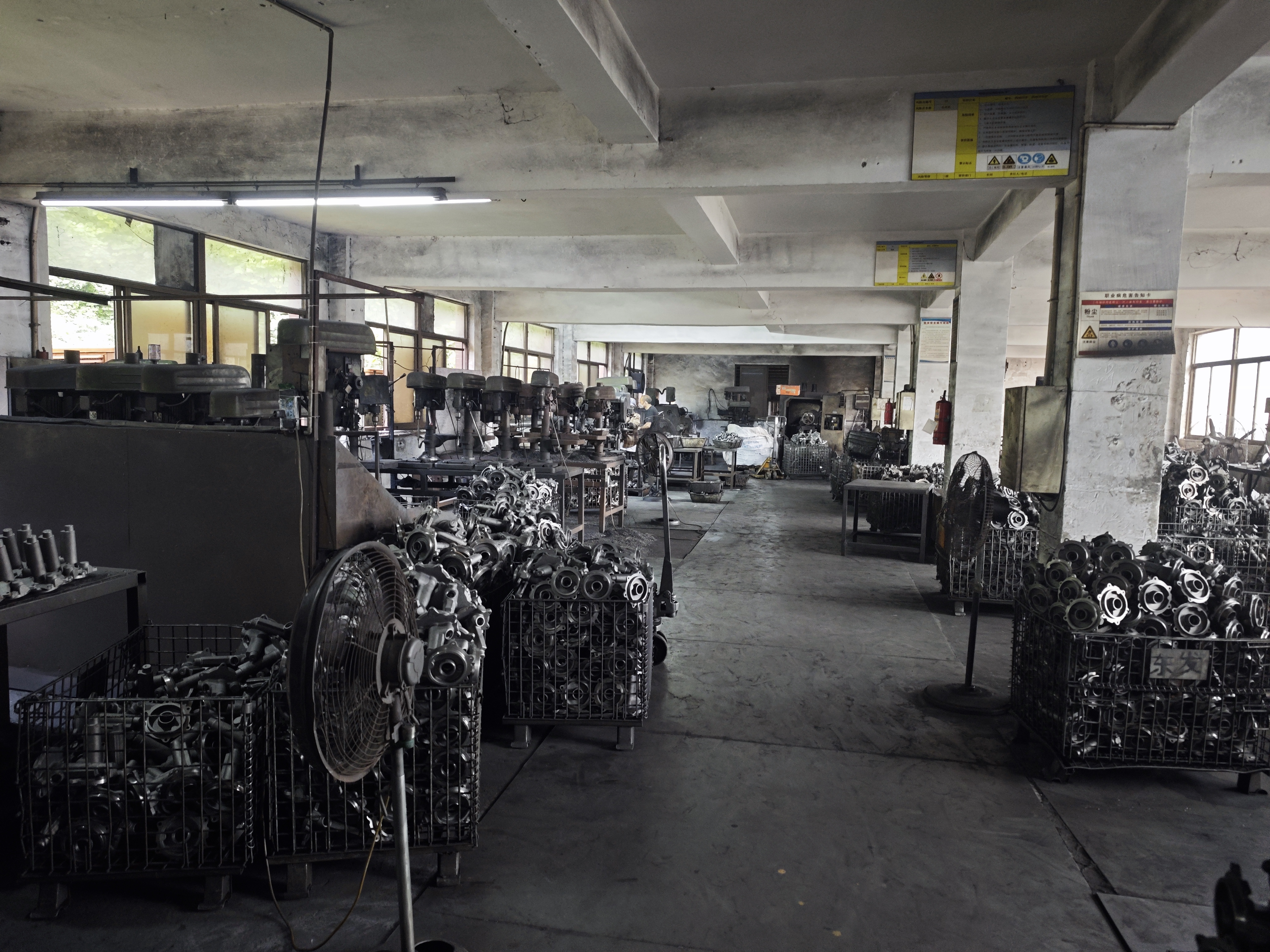 A workshop floor with a row of drill presses on the left and wire-mesh baskets full of cast housings along the aisle