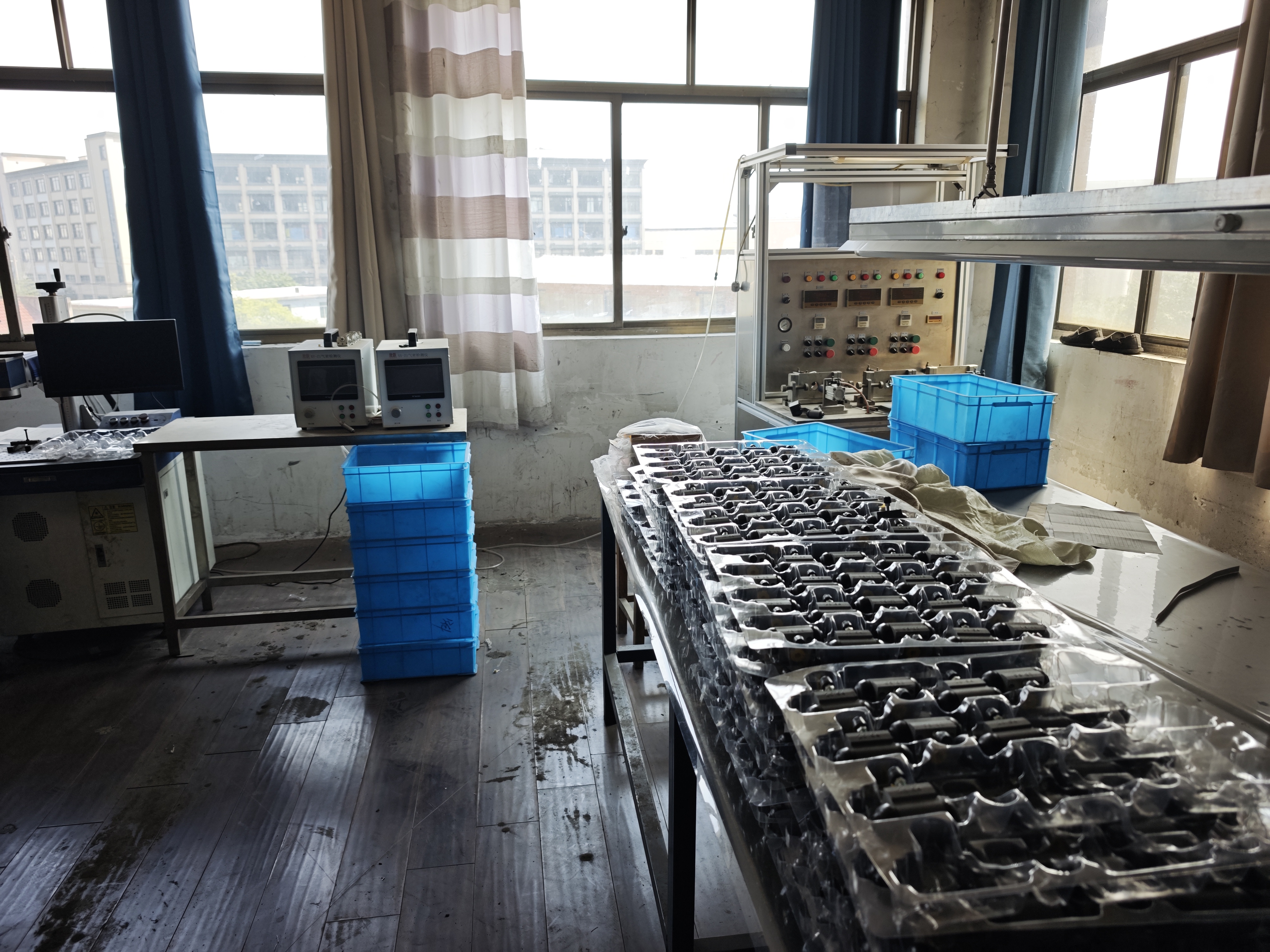 A room with two digital instrument units on a table, a control panel with gauges and switches, blue crates, and a table covered in clear blister trays holding finished parts
